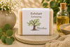 Savon exfoliant artisanal naturel La Savonnerie Blanchou présenté sur une table de bois avec huiles naturelles et feuillage au soleil